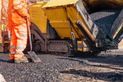 Worker in Orange Safety Gear Operating Asphalt Paving Machine on a Roadway Construction Site